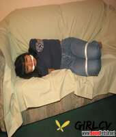 solo girl with painted toenails is hog tied & gagged on a sofa in her clothes